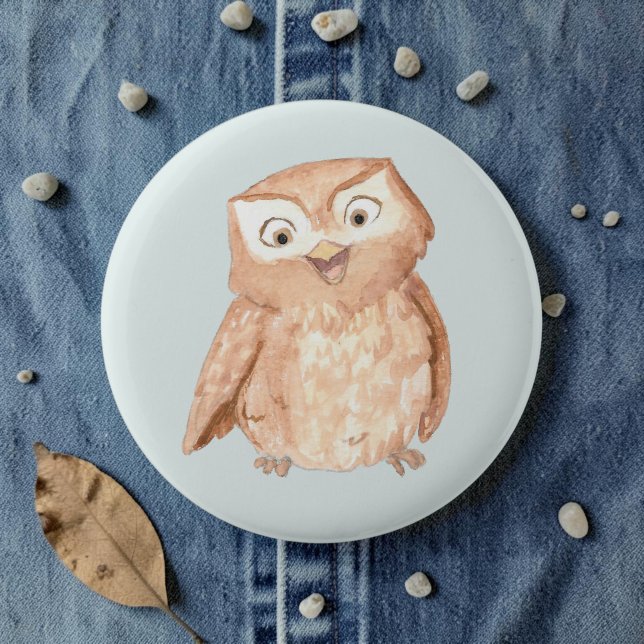 Badge Rond 5 Cm Chouette Heureuse Aquarelle (A watercolor owl cartoon smiling on a pale blue pin, laid on denim amid pebbles and leaves.)