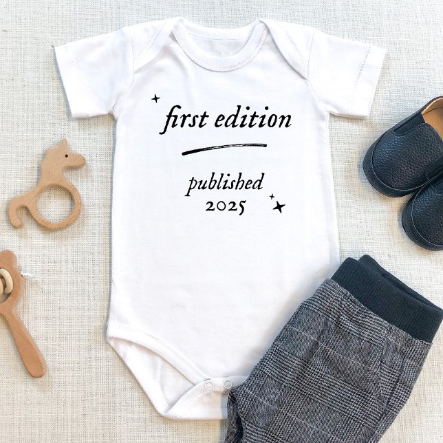 Body Première édition Livre Publié 2025 Costume pour bé (First edition baby bodysuit with personalized birth year. Shown on beige table with baby pants. book)