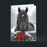 Carte Cheval de Noël<br><div class="desc">Un cheval se tient derrière une clôture en bois ornée d'une couronne festive et d'un arc rouge,  entourée d'un paysage hivernal enneigé. Le texte intérieur peut être modifié.</div>