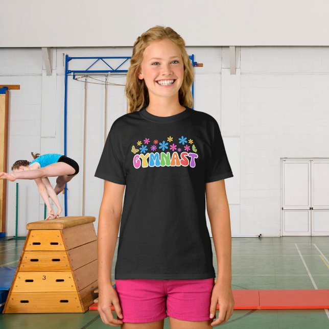 T-shirt Retro Gymnast | T-shirt à thème Gymnastiqu (A young girl wearing gymnast black t-shirt with flower power style retro design. Gymnasium behind.)