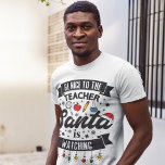 T-shirt Soyez gentil avec le professeur Père Noël regarde<br><div class="desc">Rappelez à vos élèves de rester sur la belle liste cette année avec ce t-shirt amusant avec le libellé "Soyez gentil avec l'enseignant, Père Noël regarde" en polices modernes noires, décoré de lumières de Noël rouges, vertes et jaunes, flocons de neige, chapeau de Père Noël rouge, et quelques objets scolaires...</div>
