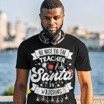T-shirt Soyez gentil avec le professeur Père Noël regarde<br><div class="desc">Rappelez à vos élèves de rester sur la belle liste cette année avec ce t-shirt amusant avec le libellé "Soyez gentil avec l'enseignant, Père Noël regarde" en polices modernes blanches, décoré avec des lumières de Noël rouges, vertes et jaunes, des flocons de neige, un chapeau de Père Noël rouge, et...</div>
