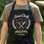 Tablier Custom Name Sous Chef Sidekick Child Apron<br><div class="desc">Make kitchen time extra fun with this personalized Sous Chef Sidekick kids apron. Featuring a stylish crest design with cooking utensils, flame detail, and custom name personalization, this apron is perfect for little helpers who love baking, cooking, and BBQ time with family. A great gift for birthdays, holidays, Father’s Day,...</div>