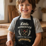 Tablier Vintage Barbecue Chef Apron Kids Apprentice<br><div class="desc">Embrace your culinary journey with the "Little Apprentice" apron, a design that blends vintage charm with modern grilling spirit. This graphic apron showcases a sophisticated emblem with a shield, featuring iconic barbecue tools and a fiery emblem, all encircled by elegant laurel branches. The phrase "Learning from the Best Since 1998"...</div>